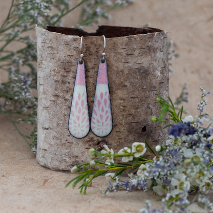 Handcrafted enamel earrings in a soft teardrop shape featuring pink and white botanical thistle-inspired patterns. Lightweight dangles with a fresh spring feel, displayed on natural birch bark with wildflowers – Spring Thistle Earrings by Divella Designs.