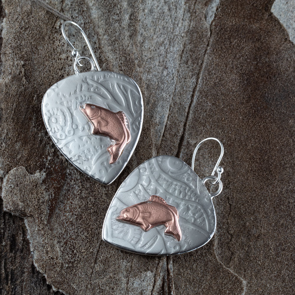 Handcrafted mixed metal earrings featuring copper jumping bass fish on textured fine silver backdrops with nature-inspired patterns – Gone Fishin’ earrings by Divella Designs, displayed on slate background.