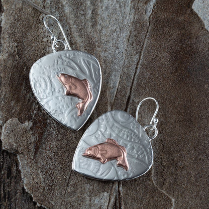 Handcrafted mixed metal earrings featuring copper jumping bass fish on textured fine silver backdrops with nature-inspired patterns – Gone Fishin’ earrings by Divella Designs, displayed on slate background.
