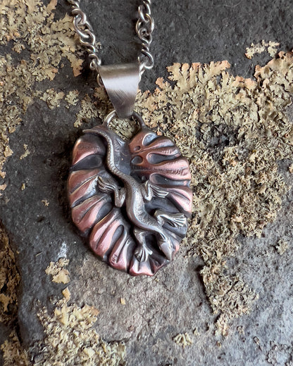 Handcrafted sterling silver and copper pendant necklace featuring a sculpted lizard on a textured leaf background, with oxidized finish and nature-inspired artisan design. Hanging on a handmade sterling chain