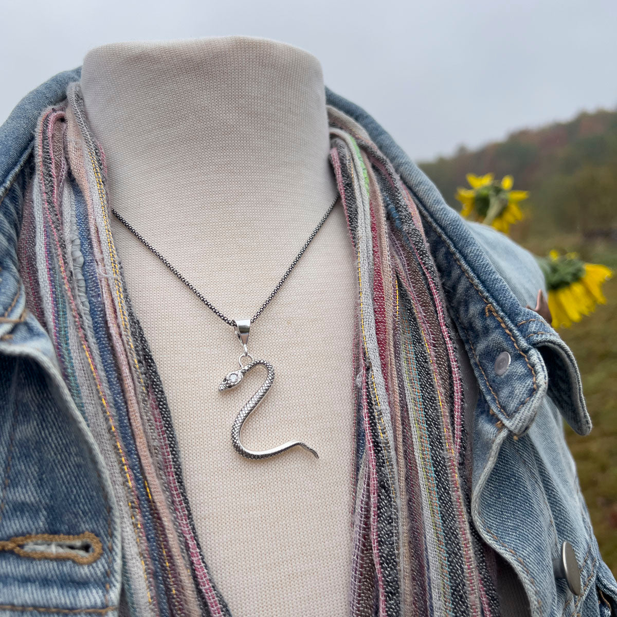 Silver swan necklace on a mannequin bust with a denim jacket and striped scarf, sunflowers in the background.