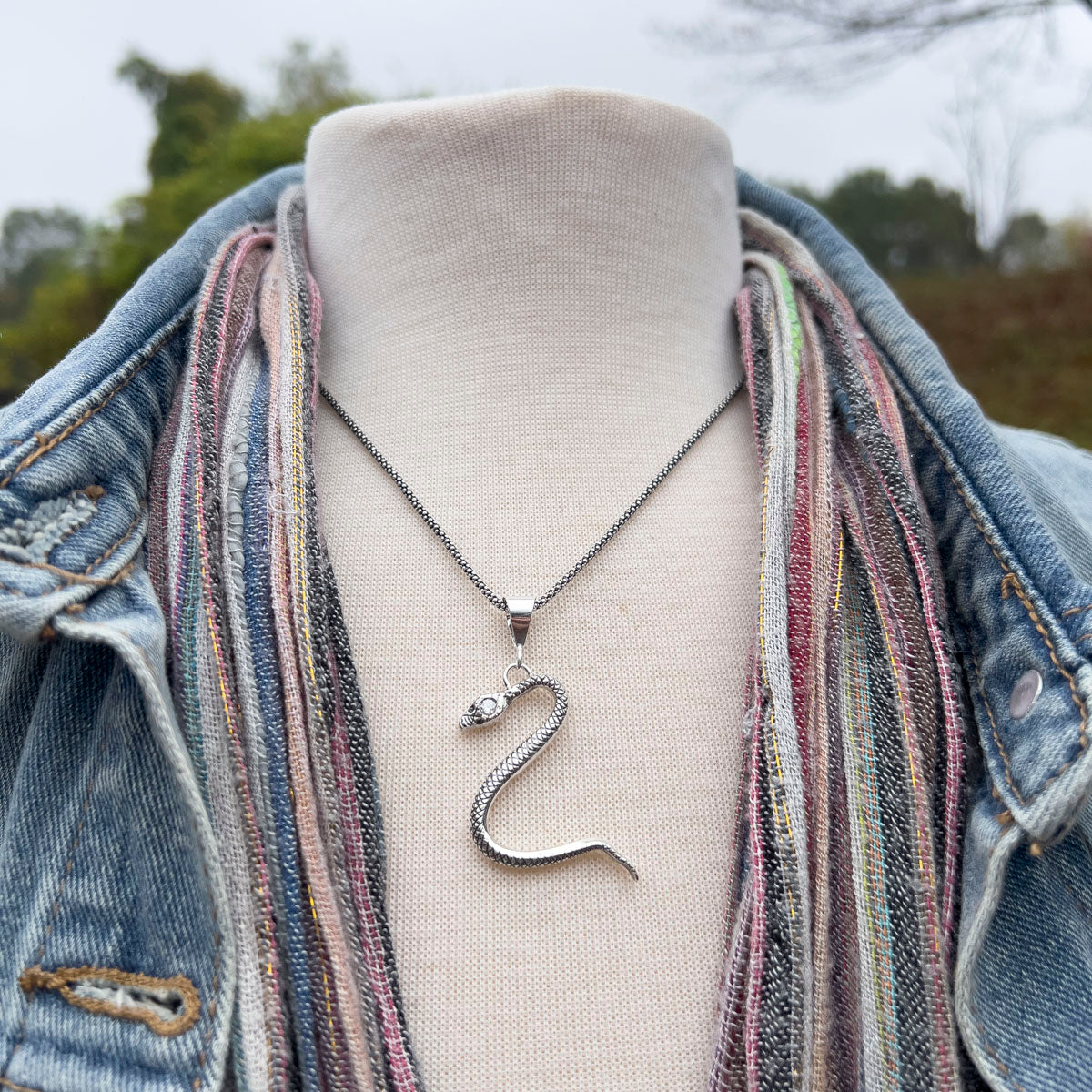 Silver snake pendant necklace on a mannequin bust with a denim jacket and colorful scarf outdoors.