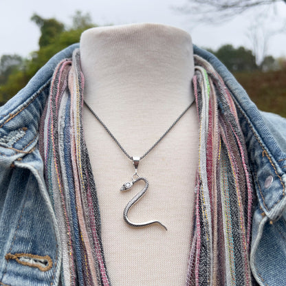 Silver snake pendant necklace on a mannequin bust with a denim jacket and colorful scarf outdoors.