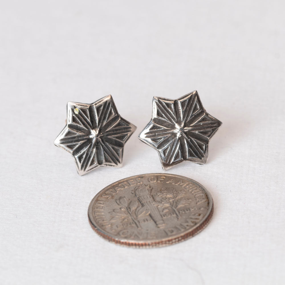 Pair of handcrafted sterling silver star-shaped stud earrings with etched radiating lines, displayed on a light fabric backgroun