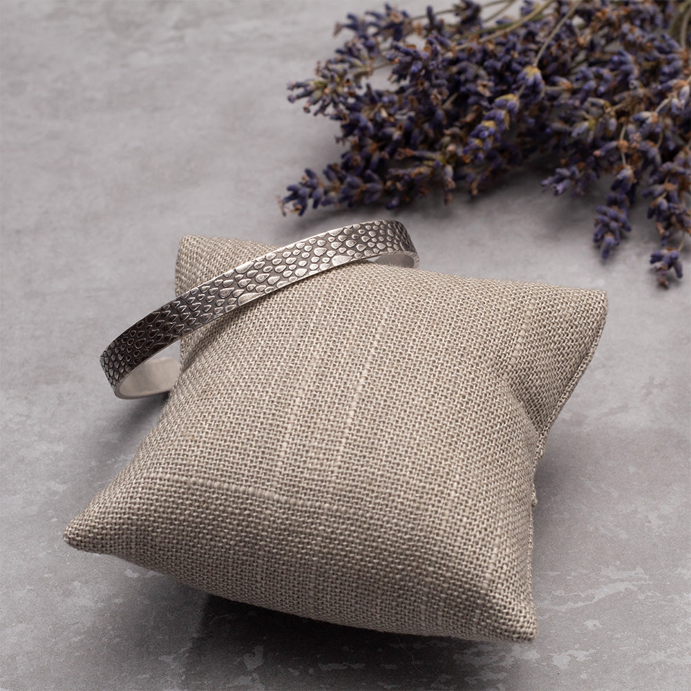 Sterling silver cuff bracelet with a raindrop texture, displayed on soft fabric pillow with dried lilac for decoration.