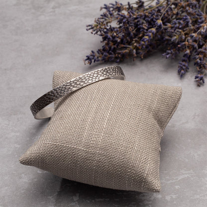 Sterling silver cuff bracelet with a raindrop texture, displayed on soft fabric pillow with dried lilac for decoration.