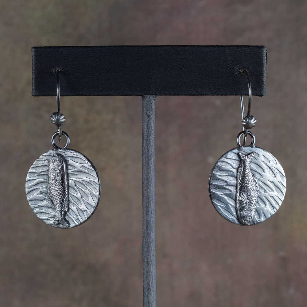 Handcrafted sterling silver earrings featuring detailed fish motifs swimming over textured water-like backgrounds – Swimmers artisan earrings by Divella Designs, displayed on earthy stone surface.