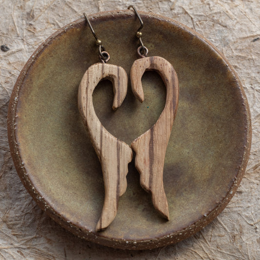 A pair of wooden dangle earrings shaped like a heart silhouette, displayed on a round rustic ceramic dish over a textured neutral surface.