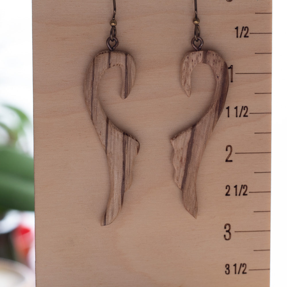 A pair of handmade wooden dangle earrings with a wavy, abstract organic shape and visible wood grain, hanging from dark metal hooks against a light wood block with measurement markings.