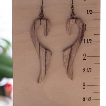A pair of handmade wooden dangle earrings with a wavy, abstract organic shape and visible wood grain, hanging from dark metal hooks against a light wood block with measurement markings.