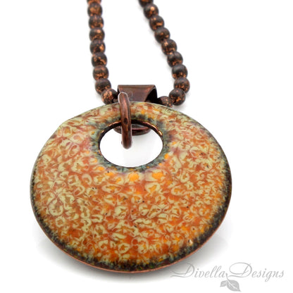 Close-up of round vitreous enamel copper pendant with earthy orange and soft green forest-inspired texture.