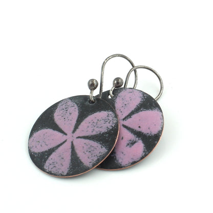 Round dangle earrings featuring a matte black background with a soft pink five-petal floral design, hanging from oxidized metal ear wires on a white background.