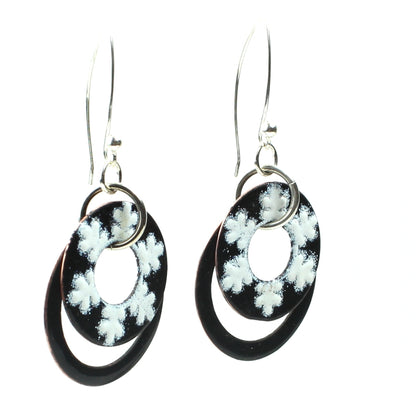 Dangling earrings with two black enamel rings, the front ring decorated with white snowflake-like patterns, hanging from silver ear wires.