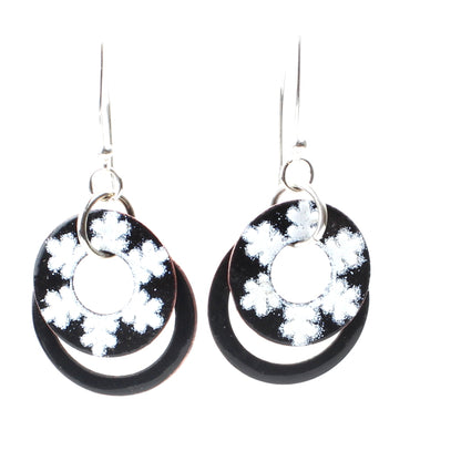 Dangling earrings with two black enamel rings, the front ring decorated with white snowflake-like patterns, hanging from silver ear wires.